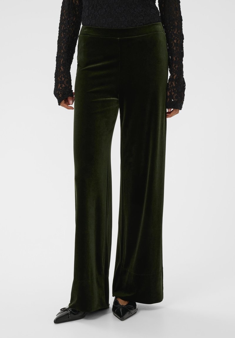 Haut en dentelle noire à manches longues associé à un pantalon en velours vert foncé à taille haute et jambes larges, accompagné de ballerines noires à bouts pointus avec des nœuds.