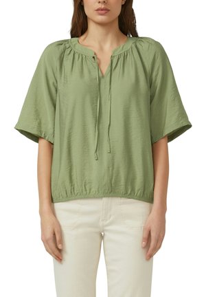 Vrouw draagt een lichtgroene blouse met korte mouwen, elastische zoom en een strik bij de hals, gecombineerd met een offwhite broek, staat met ontspannen armen.