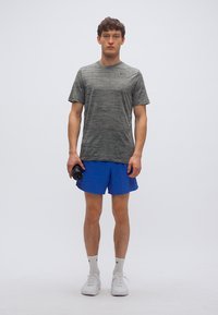 Γκρι αθλητικό κοντομάνικο με υφασμάτινη υφή, σε συνδυασμό με μπλε σορτς. Το άτομο κρατάει μια μαύρη παγούρι νερού, φοράει λευκά αθλητικά παπούτσια.