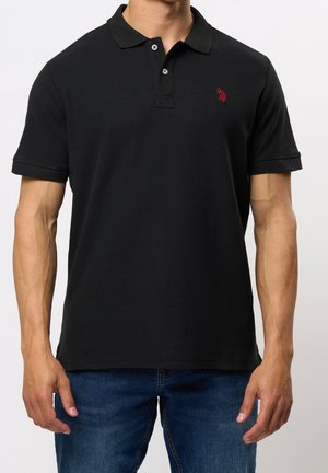 Mann trägt ein schwarzes kurzärmeliges Poloshirt mit zwei Knöpfen und einem kleinen roten Sticklogo auf der linken Brust, kombiniert mit blauen Jeans.