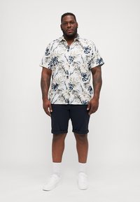 Camisa de manga corta con fondo blanco y patrón de hojas en azul y verde. Combinada con pantalones cortos azul marino y zapatillas blancas, completada con calcetines blancos.