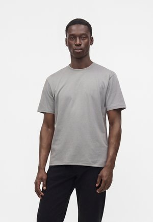 Junger Mann mit kurzen Haaren, der ein einfarbiges hellgraues kurzärmeliges T-Shirt und schwarze Hose trägt, steht vor einem weißen Hintergrund.