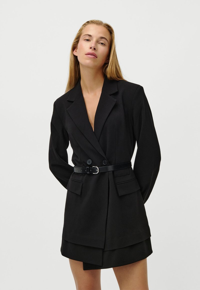 Kvinde med langt blond hår iført en sort blazer-kjole med bælte, revers og klaplommer, poserende mod en ensartet hvid baggrund.