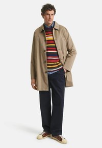 Beige, lichte trenchcoat met een knopenfront. Daaronder een multikleurige gestreepte trui, marineblauwe broek en crème- en bruine sneakers.