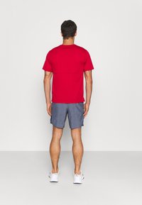 Rode shirt met korte mouwen en een ronde hals, gecombineerd met grijze shorts. De outfit wordt aangevuld met witte sportschoenen.