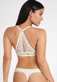Kant bralette in crème met een driehoekig rugontwerp, verstelbare bandjes en gegolfde randen. Bijpassende kanten string inbegrepen.