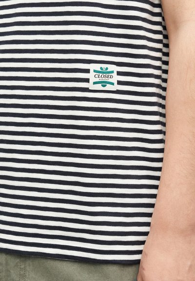 Κοντινή λήψη μιας μαύρης και άσπρης ριγωτής μπλούζας με μικρό λογότυπο «Closed Workshop» κοντά στο τελείωμα, δίπλα σε ένα χέρι και πράσινο παντελόνι.