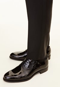 Schwarze Lackleder-Absatzschuhe mit runder Zehenform, Schnürung im oberen Bereich und einer polierten Oberfläche. Kombiniert mit schmal geschnittenen schwarzen Hosen.