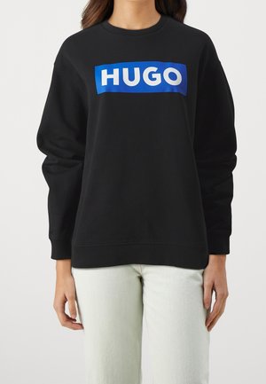 Osoba nosząca czarną bluzę z niebieskim kwadratem i białym napisem "HUGO", w połączeniu z jasnymi spodniami, stojąca na jednolitym tle.