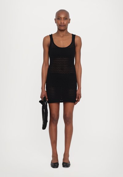 Rochie mini tricotată neagră, cu o siluetă ajustată, având un decolteu circular și un model din plasă deschisă. Modelul ține o geantă neagră asortată.