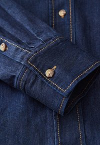 Polsino della camicia in denim di colore blu scuro con cuciture visibili. Presenta bottoni beige e una texture liscia, mettendo in evidenza la durabilità e il design classico.