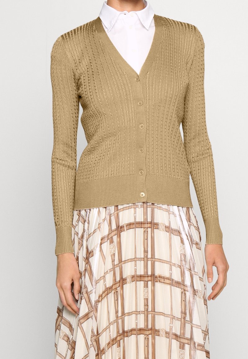Pletený kardigan v béžové barvě s žebrovanou texturou, s výstřihem do V, dlouhými rukávy a knoflíky vpředu. Nosič z něj obléká vzorovanou sukni.
