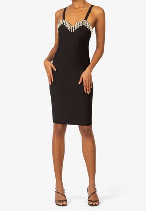 Femme portant une robe noire ajustée sans manches avec un détail de franges perlées le long de l'encolure en cœur et des talons noirs à brides.