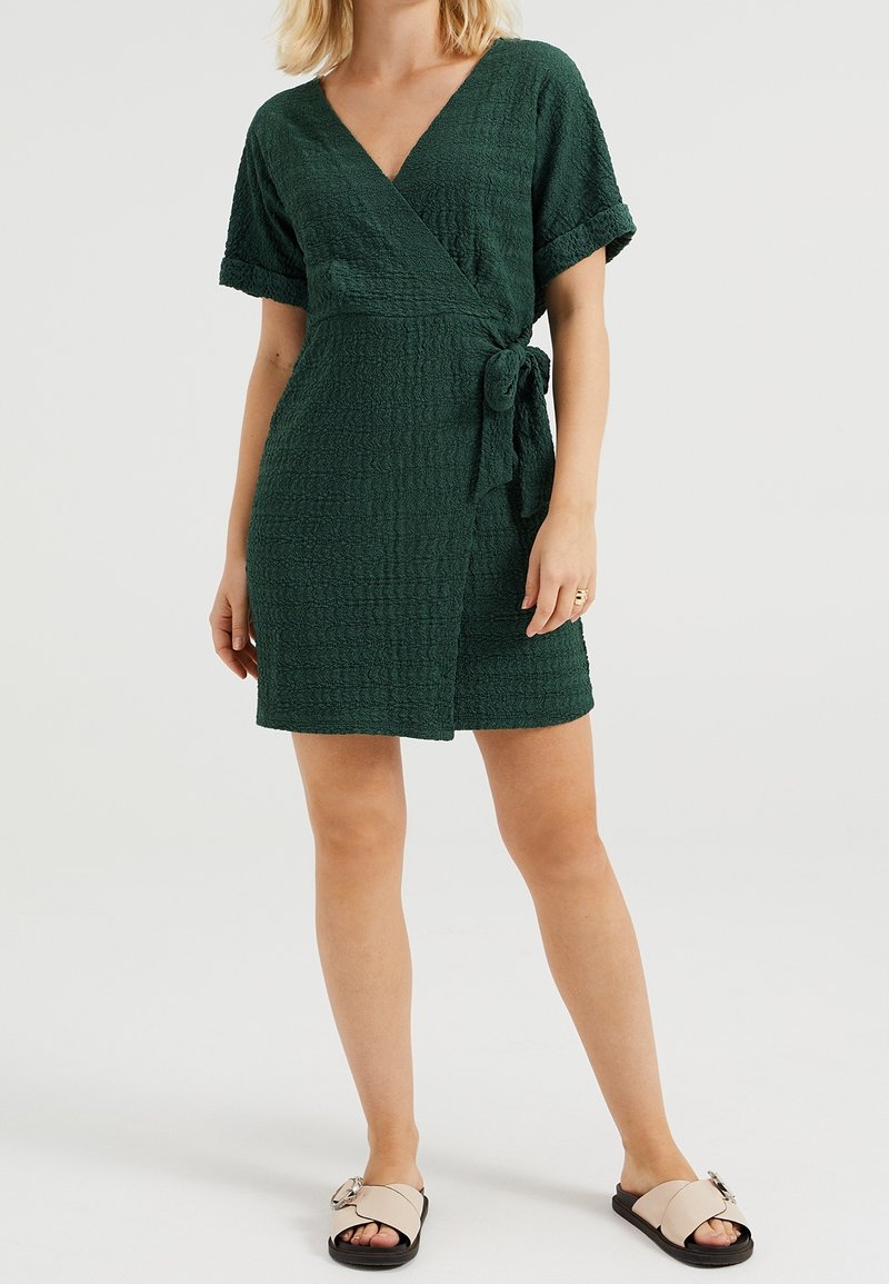 Robe d'enveloppe verte texturée avec des manches courtes, nouée à la taille, présentant un décolleté en V profond. Associée à des sandales beiges.