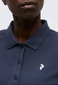 Gros plan d'une personne portant un polo bleu marine avec un col, trois boutons et un petit logo blanc sur la poitrine.