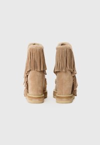 Botas de tobillo de ante en beige claro con detalles de flecos en la parte posterior. Cuenta con un suave collar redondeado y una suela de plataforma baja y texturizada.