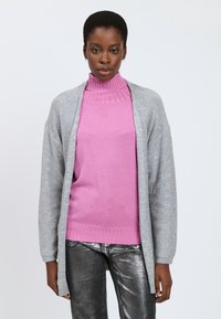 Cárdigan gris de corte holgado con detalles brillantes sobre un suéter de cuello alto rosa. Jeans ajustados metálicos plateados completan el conjunto.