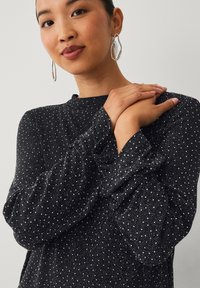 Zwarte top met lange mouwen van een gestructureerde stof met zilveren spikkels. Model draagt oversized creolen en heeft de armen gekruist voor het lichaam.