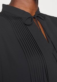 Blouse noire avec col montant à nouer, texture plissée sur le devant et ouverture en forme de goutte d'eau au niveau de l'encolure.