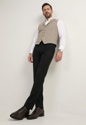 Mand, der læner sig op ad en væg iført en beige tekstureret vest, hvid skjorte, sorte bukser, brune pæne sko og et sølvfarvet armbåndsur.