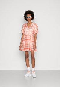 Vestido curto xadrez em tons de rosa e laranja, com mangas bufantes, decote em V e cintura atada. Combinado com sapatilhas brancas de plataforma.