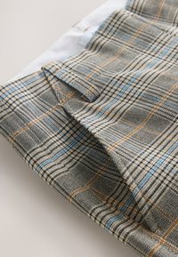 Primer plano de tela gris con cuadros de líneas azules y marrones doblada, mostrando una cinturilla y detalle de bolsillo lateral sobre una superficie blanca.