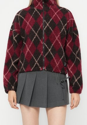 Femme portant une veste polaire zippée à motif losanges rouge et noir et une mini-jupe plissée grise avec un détail de nœud sur le côté.