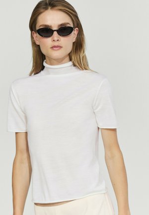 Femme portant des lunettes de soleil ovales noires et un haut blanc à manches courtes et col montant sur un fond clair.