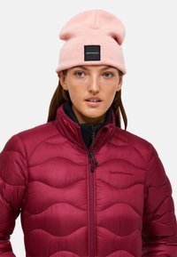 Bordeaux kleurige pufferjack met een gewatteerd patroon, voorzien van een hoge kraag en ritssluiting. Roze gebreide beanie met een zwart label aan de voorkant.