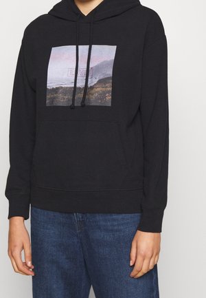 Personne portant un sweat à capuche noir avec un motif de paysage montagneux et le logo "Levi's", associé à un jean bleu.