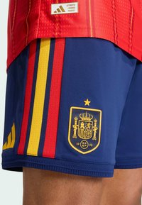 Červený dres se žlutými a červenými pruhy, doplněný modrými šortkami s zlatým znakem Španělska a logem Adidas. Hladká tkanina, atletický střih.