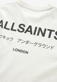 Bílý bavlněný tričko s výrazným černým textem: "ALLSAINTS," japonské znaky a "LONDON" na spodní části. Hladká textura, ležérní design.