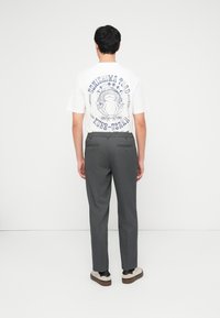 Άνδρας με πλάτη φορούσε λευκό μπλουζάκι με γραφικό βάτραχο και φεγγάρι και γκρι παντελόνια, στέκεται μπροστά σε απλό λευκό φόντο.