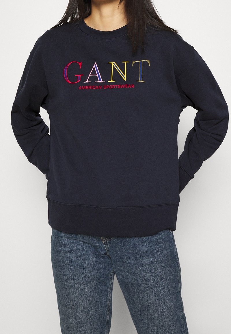 Felpa blu navy con scollatura tonda, polsini e orlo a coste. Presenta il testo ricamato multicolore "GANT AMERICAN SPORTSWEAR" sul davanti.
