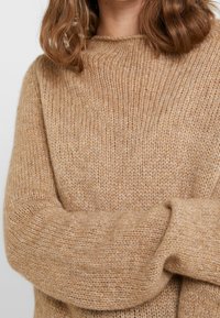 Sweter dziany w jasnym beżu, o luźnym kroju, okrągłym dekolcie i złożonych mankietach; teksturowany, miękki materiał z delikatnym wzorem.