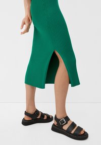 Robe midi verte à côtes avec une fente sur le côté, associée à des sandales en cuir noir à plusieurs brides avec un détail à boucle.