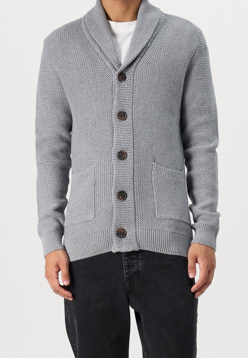 Hombre vistiendo un cárdigan de punto con cuello chal en gris claro, con botones oscuros y dos bolsillos frontales, sobre una camisa blanca y pantalones negros.