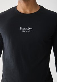 Black long-sleeve t-shirt made of cotton. Features white embroidered text "Brooklyn NEW YORK" at the chest. Simple, fitted design.