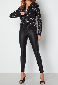 Veste bomber noire à motifs floraux avec une fermeture éclair à l'avant, des poignets et un ourlet côtelés. Associée à des leggings noirs moulants et brillants et à des talons noirs ouverts.