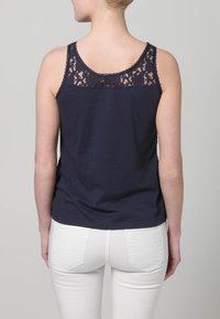 Granatowy top na ramiączkach z koronkowymi detalami na plecach, wykonany z miękkiej bawełny. Noszony z białymi dżinsami. Prosty fason, bez rękawów.