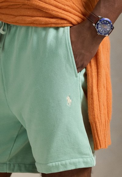 Világoszöld pamut rövidnadrág kis fehér logó hímzéssel a bal lábszáron, narancssárga kötött pulóverrel párosítva. Barna óra a csuklón.