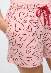 Ροζ σορτς από μαλακό ύφασμα με επαναλαμβανόμενο μοτίβο καρδιών σε σκούρο κόκκινο. Διαθέτουν ελαστική μέση με σούρα και τσέπες.