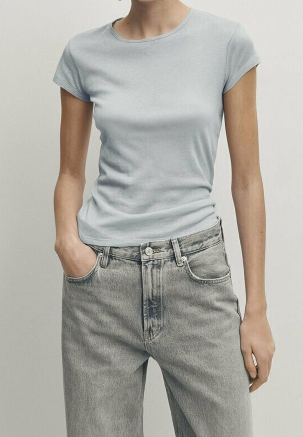 Femme portant un t-shirt ajusté à manches courtes bleu clair et un jean gris délavé taille haute, avec une main dans la poche, debout devant un fond uni.
