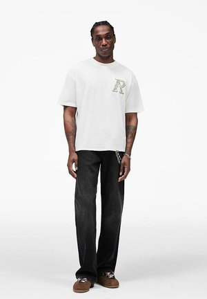 Man staand, gekleed in een oversized wit T-shirt met een klein ontwerp op de borst, zwarte wijduitlopende jeans en bruine schoenen met patroon, tegen een effen witte achtergrond.