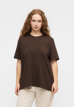 Νέα γυναίκα με κυματιστά κόκκινα μαλλιά που φορά χαλαρό σκούρο καφέ μπλουζάκι και ανοιχτά μπεζ παντελόνια, στέκεται μπροστά σε ένα απλό ανοιχτό φόντο.