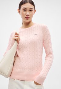 Svetloružový sveter s vrkočovým vzorom, okrúhlym výstrihom, rebrovanými manžetami a lemom. S malým logom na hrudi. Hladká textúra.