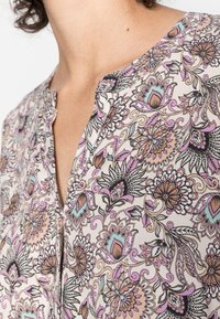 Blusa floral com um fundo bege claro, apresentada com padrões florais em roxo, teal e preto; decote em forma de olho-chave; tecido leve e suave.
