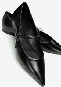 Par de zapatos Mary Jane negros con punta puntiaguda, con una correa fina y un pequeño detalle de lazo sobre un fondo blanco.