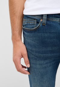 Vīrieša roku balstās pie zilo džinsu kabatas, uzvilktā baltā krekla fons uz vienkārša gaiša fona.