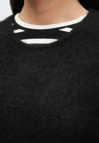 Juodas, pūkuotas megztinis su apvalia iškirpte ir sluoksniuotu baltos ir juodos dryžuotos vidinės marškinėlių dalies, turinčios iškirpimus.